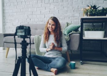 Happy female blogger is recording video for internet videolog talking and gesturing showing thumbs-up and waving hand. Blogging, young people and success concept.