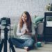 Happy female blogger is recording video for internet videolog talking and gesturing showing thumbs-up and waving hand. Blogging, young people and success concept.