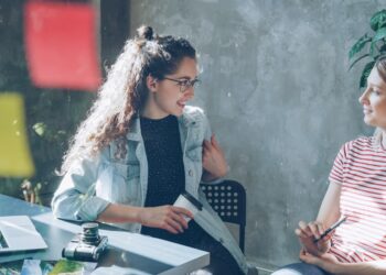 Businesswomen are sitting at desk together and discussing new project in modern office indoors. Glass with colourful stickers in foreground.