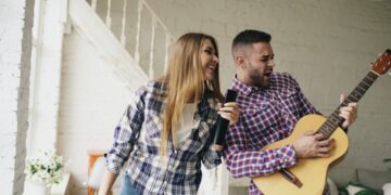 Funny happy and loving couple dance on bed singing with tv controller and playing guitar. Man and woman have fun during holiday at home